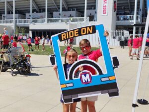 Two people posing in front of Energizer Park with a MetroBus-shaped prop that says "RIDE ON"